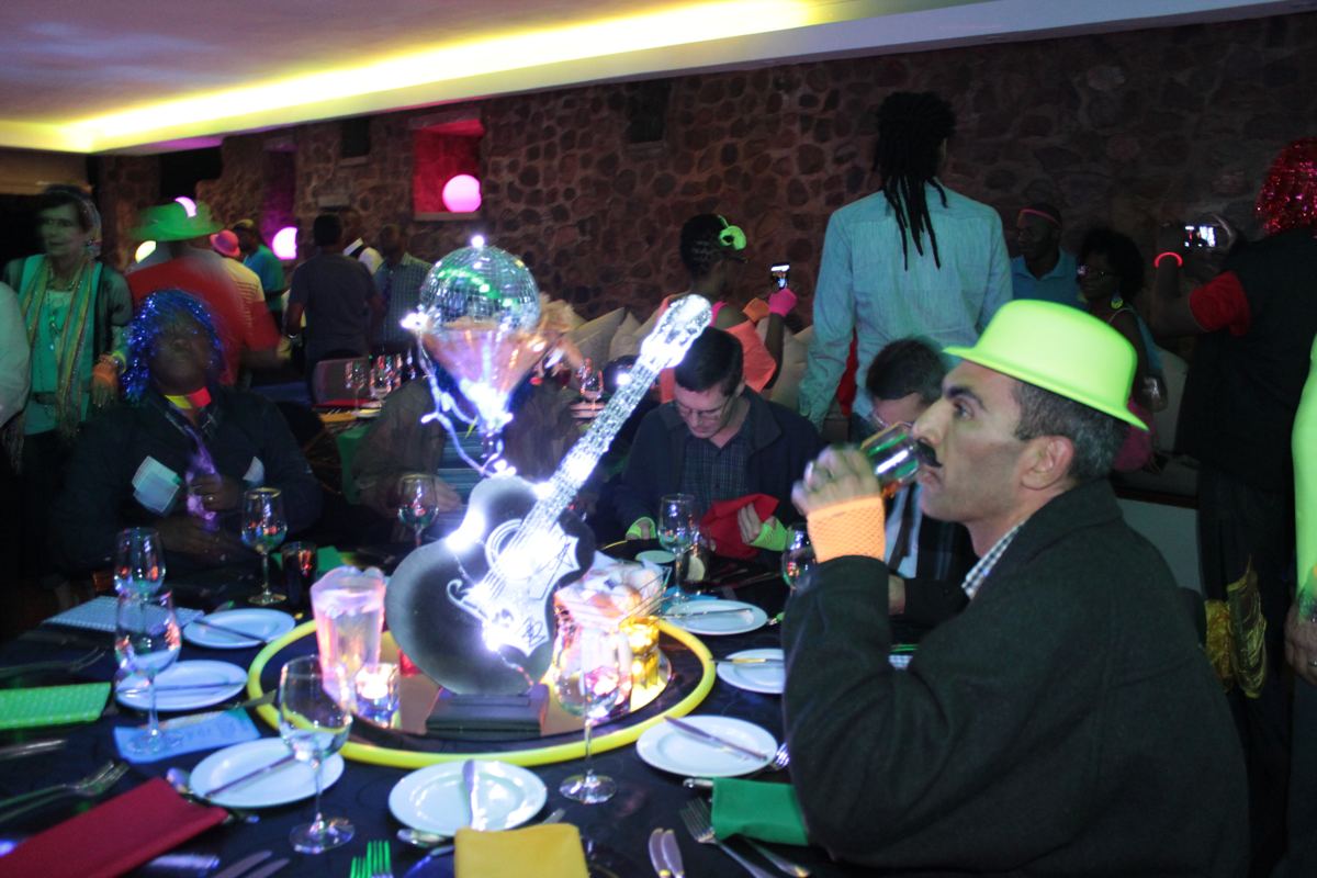 Guests in neon hats and 80s outfits at a themed year-end function dinner with lit guitar and disco ball centrepieces