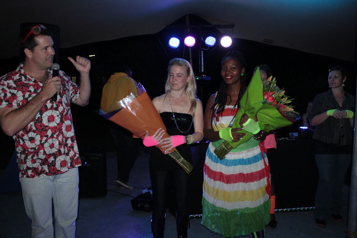MC presenting flower bouquets to two staff award winners at an 80s themed year-end function